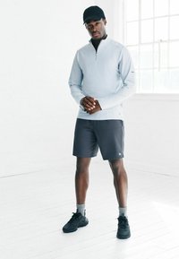 Vīrietis ar melnu cepuri, gaiši pelēku garām piedurknēm zip-up krekli, tumši pelēkiem šortiem, pelēkām zeķēm un melnām sporta apaviem stāv spilgti baltā istabā.