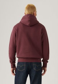 Burgundy hoodie with a hood, ribbed cuffs, and waistband, paired with dark blue jeans showcasing a classic back pocket design.