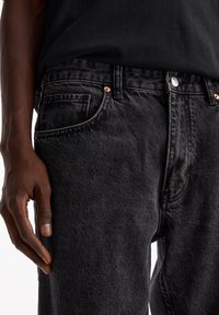 Jeans in denim nero con una texture sbiadita, dotati di un design a cinque tasche, rivetti in rame e chiusura con bottoni. La mano del modello poggiata sulla cintura.