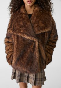 Bruine nepbontjas met een brede kraag, gestructureerd oppervlak en oversized mouwen. Gecombineerd met een geruite rok in meerdere bruin- en beigetinten.