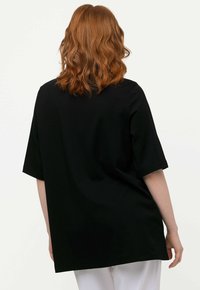 Top nero a maniche corte con vestibilità rilassata, realizzato in un tessuto morbido; presenta un colletto rotondo e scende sotto i fianchi. La parte posteriore è semplice e priva di decorazioni.