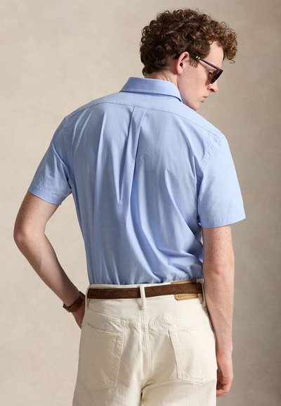 Homme aux cheveux bouclés portant des lunettes de soleil, une chemise bleu clair à manches courtes rentrée dans un pantalon blanc avec une ceinture marron, vu de dos.