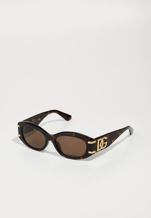 Óculos de sol de padrão tartaruga com lentes castanhas, de forma oval, armação espessa e detalhes dourados com um logótipo proeminente de um lado.