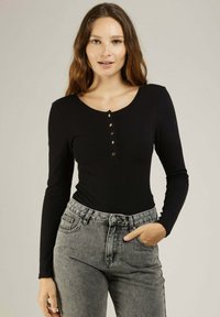 Top noir à manches longues avec un col rond, présentant une rangée de boutons métalliques à l'avant. Associé à un jean gris délavé taille haute.