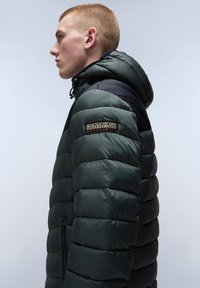 Giacca puffer imbottita di colore verde scuro con un pannello nero sulla spalla, con una toppa logo sulla manica e tasche laterali con zip.