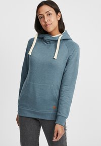 Sudadera con capucha azul y cordones color crema, con un bolsillo frontal, puños de canalé y un parche de logotipo en la esquina inferior izquierda; textura de tela suave.