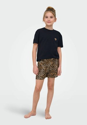 Dziewczyna stoi boso, ubrana w czarną koszulkę z małą łatką w panterkę oraz krótkie spodenki w panterkę na jednolitym białym tle.