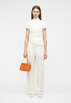 Femme portant un haut blanc côtelé à manches courtes et un pantalon à jambes larges, tenant un sac à main orange avec une sangle chaîne dorée.