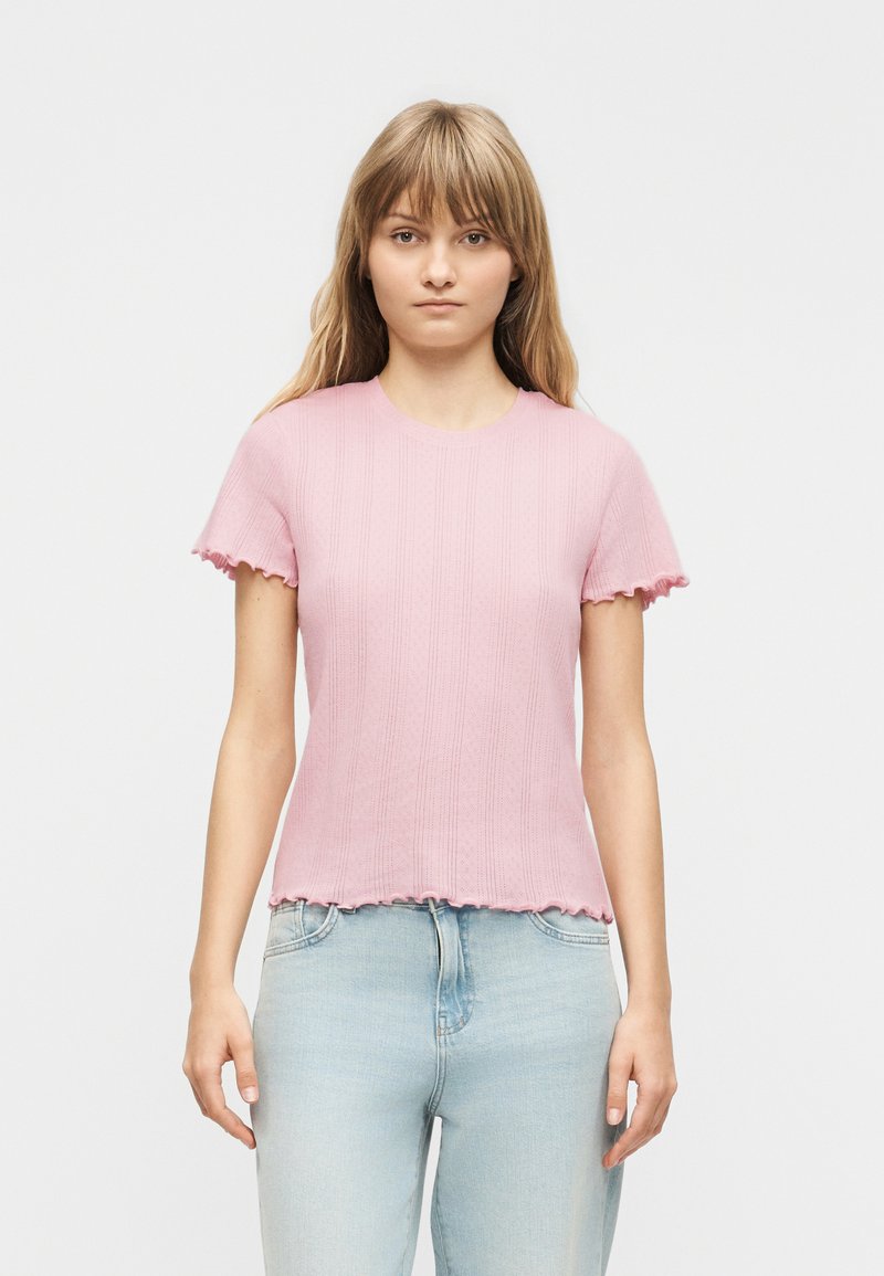 Νεαρή γυναίκα με ίσια ξανθά μαλλιά φοράει ελαφρύ ροζ ριμπ κοντομάνικο τοπ και ανοιχτό μπλε τζιν, στέκεται μπροστά σε μια απλή φόντο.