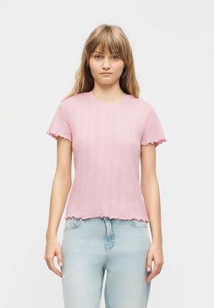 Mladá žena s rovnými blond vlasy má na sobě světle růžový žebrovaný krátký rukáv a světlé modré džíny, stojí před jednobarevným pozadím.