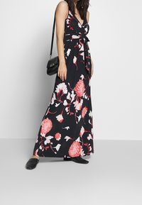 Maxi šaty v černé tkanině s velkými růžovými a bílými květinovými vzory, s tkaničkou v pase a tenkými ramínky. Černá taška a ploché boty.