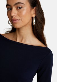 Top azul marino de hombros descubiertos con textura acanalada, que cuenta con un escote amplio. La modelo lleva pendientes hoop plateados brillantes para un detalle adicional.