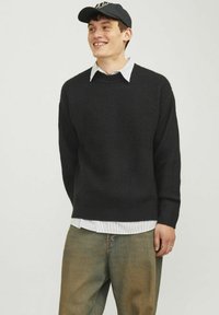 Schwarzer gestrickter Pullover mit strukturiertem Muster, kombiniert mit einem weißen, gestreiften Hemd mit Kragen. Getragen mit lockeren Jeanshosen und einer Mütze.