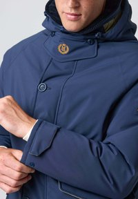 Marine vandtæt jakke med høj krave, der har et guld logo, knaplukning foran og kontrasterende mansjetter. Glat tekstur med en tætsiddende design.