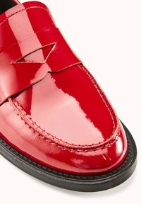 Mocassin en cuir rouge brillant avec un design cousu, présentant un accent décoratif découpé sur le dessus et une semelle en caoutchouc noire.