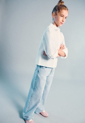 Mädchen mit blondem Haar in einem Dutt, trägt eine weiße Bluse, hellblaue Jeans und rosa Schuhe, steht mit verschränkten Armen vor einem grauen Hintergrund.