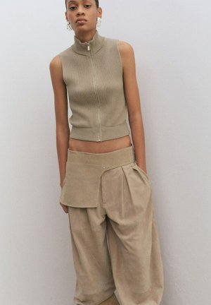 Mujer vestida con top sin mangas beige acanalado con cremallera y pantalones beige sueltos con un bolsillo con solapa asimétrica, de pie frente a una pared lisa y clara.