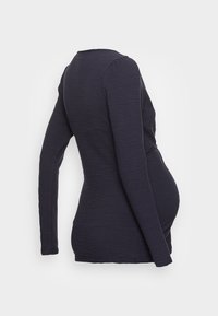 Marinblå långärmad graviditetstopp med texturerad tyg, figurnära design och lätt rynkning över magen, visad från en sidobakvinkel.