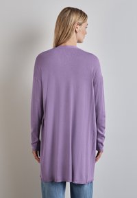 Cardigan à manches longues, ample, en tissu lilas doux. Il présente un ourlet arrondi et des fentes sur les côtés, conçu pour le confort et la liberté de mouvement.