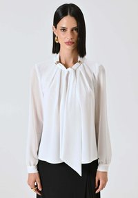 Camicia bianca con davanti drappeggiato, fiocchi al collo, maniche lunghe e dettagli in tonalità oro. Tessuto liscio con vestibilità morbida, abbinata a una gonna nera.