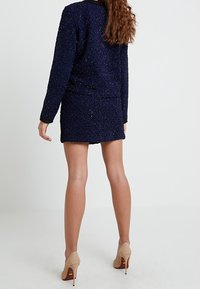 Tenue bleu marine comprenant un haut texturé et à motifs ainsi qu'une mini-jupe assortie. Des escarpins beiges unis complètent le look mais ne font pas partie de la tenue.