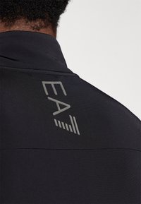 Crna sportska jakna s glatkom teksturom i visokim ovratnikom. "EA" logo u sivoj boji na poleđini, s suptilnim detaljima šivanja.