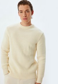 Cremfarbener, gerippter Strickpullover mit Rundhalsausschnitt und langen Ärmeln, der über einen strukturierten Stoff und einen taillierten Saum verfügt.