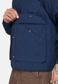 Marinblå quiltad jacka med en stor klaffficka, metallknappar och en diskret broderad logotyp på den nedre fickan.