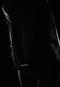 Persona vestida con una chaqueta con capucha negra, con puntos reflectantes en la manga y una pequeña franja reflectante cerca de la cintura, contra un fondo oscuro.