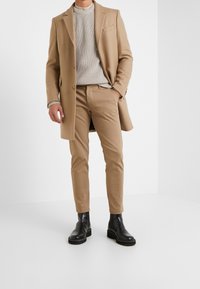 Mężczyzna w beżowym płaszczu, beżowych spodniach, szarym swetrze z dzianiny oraz czarnych skórzanych botkach Chelsea, stojący na białym tle.