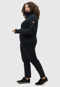 Maglione nero con dettagli a motivo, colletto alto e polsini a coste, abbinato a jeans neri e sneakers nere, evidenziando un look casual.