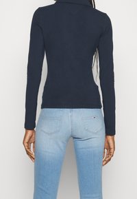 Person in einem navyblauen Langarmoberteil und hellblauen Denim-Jeans steht mit dem Rücken zur Kamera vor einem neutralen Hintergrund.