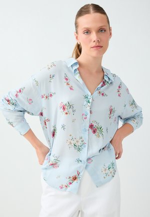 Femme aux cheveux châtain clair attachés en queue de cheval, portant une chemise bleu clair boutonnée avec des imprimés floraux multicolores et un pantalon blanc, les mains sur les hanches.
