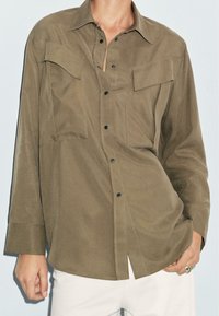 Olive green shirt wykonany z gładkiego materiału, z dwiema kieszeniami na piersi, zapinany na guziki z przodu oraz długimi rękawami w luźnym kroju.