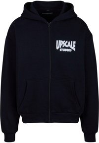Haut à capuche zippé noir en matériau polaire. Il présente une poche kangourou sur le devant et un texte logo imprimé en blanc "UPSCALE STUDIOS" sur la poitrine.