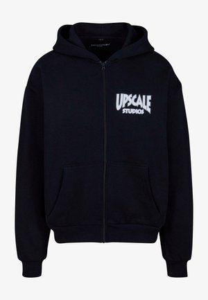 Čierna mikina na zips vyrobená z fleecového materiálu. Obsahuje predné klokanie vrecko a biely potlačený text "UPSCALE STUDIOS" na hrudi.