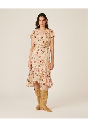 Mujer con un vestido cruzado beige con estampado floral, volantes y cinturón, combinado con botas vaqueras de ante marrón claro, de pie frente a un fondo liso.