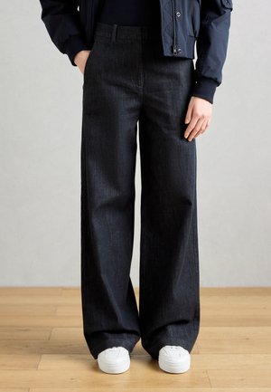 Personne portant un pantalon ample gris foncé à jambes larges, des baskets blanches, un haut noir et une veste bleu marine, mains le long du corps sur un sol en bois.
