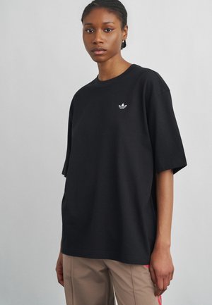 Młoda osoba ubrana w luźną czarną koszulkę Adidas z logo na piersi i beżowe spodnie, stojąca na jednolitym jasnoszarym tle.