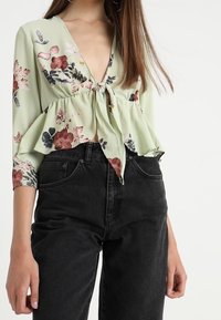 Lys grøn peplum-top med blomstermønster i pink, rød og grøn. Har en dyb V-hals og bundet front. Stylet med sorte højtaljede jeans.
