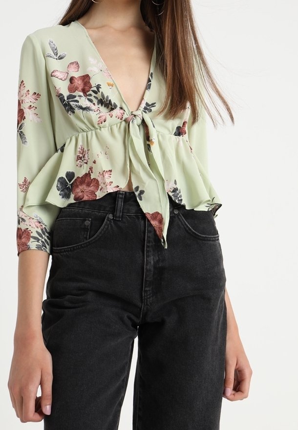Lys grøn peplum-top med blomstermønster i pink, rød og grøn. Har en dyb V-hals og bundet front. Stylet med sorte højtaljede jeans.
