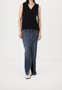 Femme portant un haut noir sans manches à col en V, une longue jupe en jean avec une fente à l'avant, et des chaussures blanches à bout pointu, sur fond blanc.