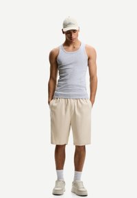 Sivý tank top, béžové šortky, béžové tenisky s bielymi podrážkami, v kombinácii s svetlou šiltovkou s textom. Model stojí s rukami v vreckách.