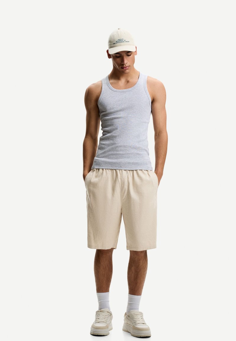 Sivý tank top, béžové šortky, béžové tenisky s bielymi podrážkami, v kombinácii s svetlou šiltovkou s textom. Model stojí s rukami v vreckách.
