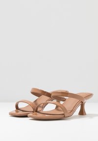 Tanfärgade klacksandaler har en glansig remdesign med två breda band som korsar foten och en avsmalnad klack. Slät läderliknande yta.