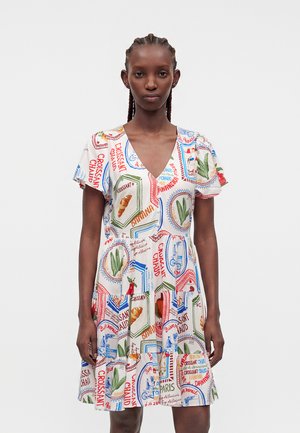 Femme noire aux cheveux tressés portant une robe blanche à col en V avec des motifs colorés de nourriture française et de texte, debout devant un fond uni.