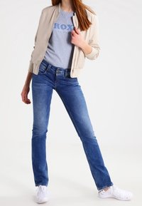 Beige bomberjacka över en grå t-shirt med blå text, kombinerad med blå jeans och vita sneakers. Utseendet har en avslappnad passform och avslappnad stil.