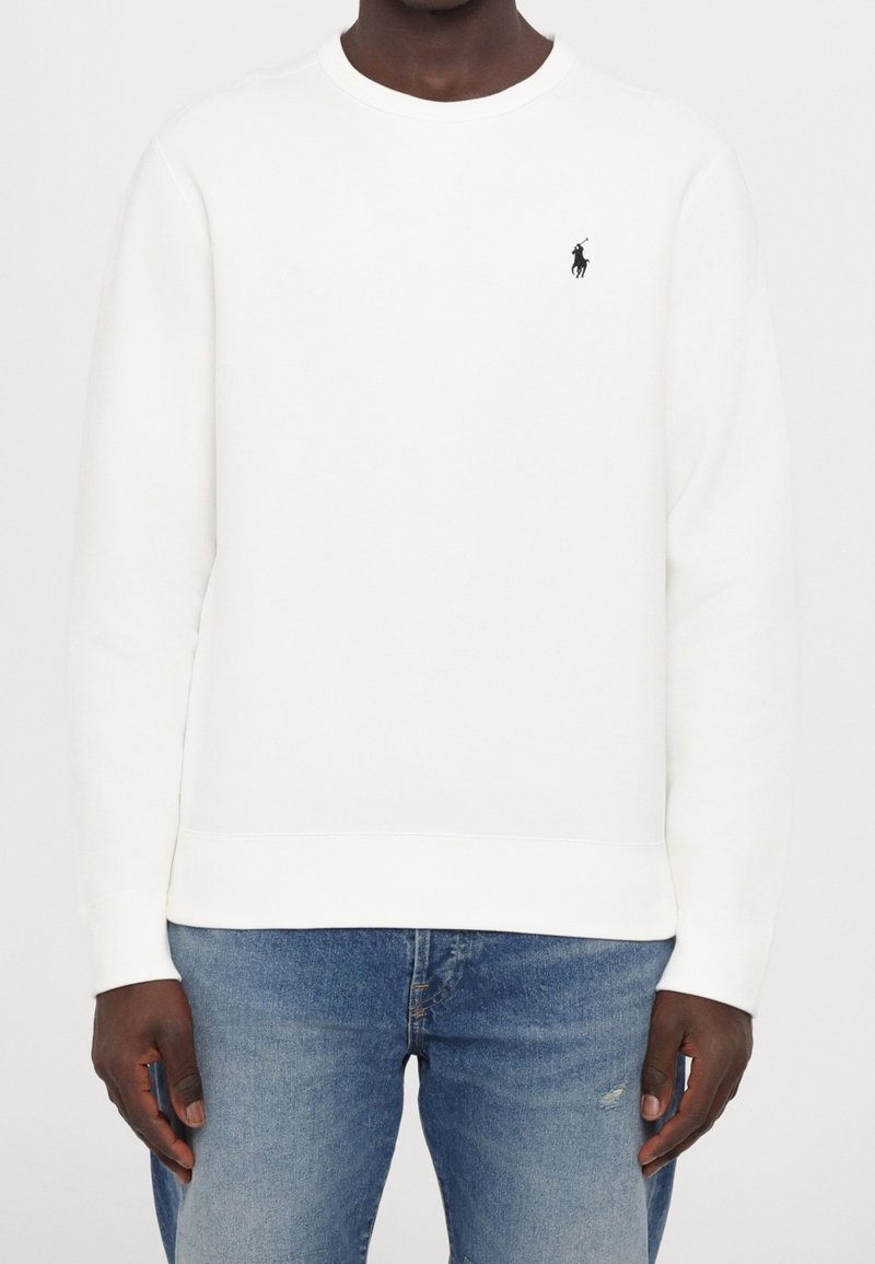 Homme portant un sweat-shirt blanc uni à manches longues avec un petit logo noir de joueur de polo sur la poitrine gauche, assorti avec un jean bleu.