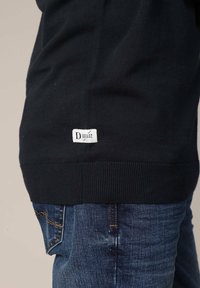 Sweat-shirt noir en tissu tricoté doux, doté d'un ourlet côtelé et d'une petite étiquette blanche avec l'inscription "Deluxe Original" près du bas. Jean bleu visible.