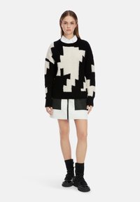 Schwarzer und weißer übergroßer Pullover mit strukturiertem Muster, kombiniert mit einem kurzen weißen gesteppte Rock mit schwarzen Akzenten und einem Reißverschluss.